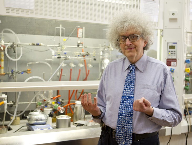 Martyn Poliakoff standing in front of fume cupboard with a series of tubes in it. He is wearing safety specs, a light blue shirt, and a brighter blue tie with the periodic table on.