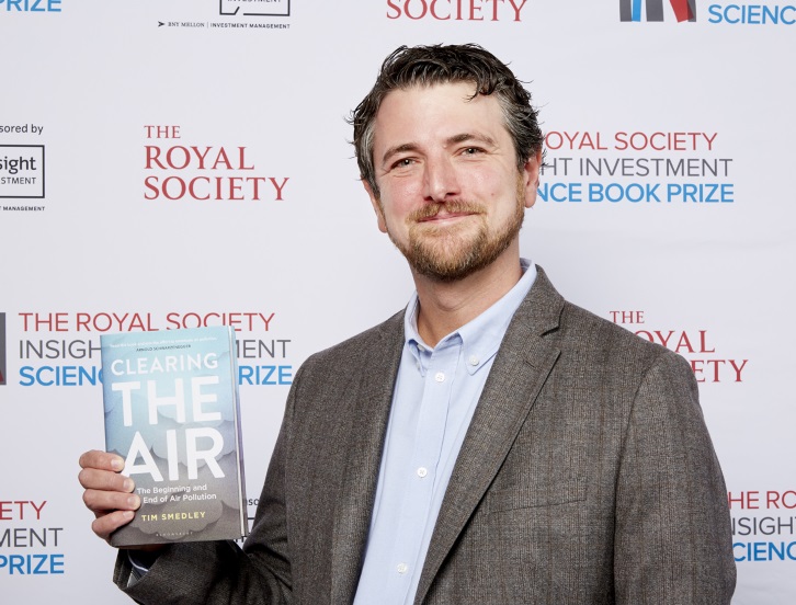 Tim Smedley wearing light blue shirt with grey suit jacket holding the book, Clearing the Air, which has a light blue cover that fades to grey at the bottom of the book cover